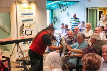 Auf diesem Bild sind viele Heimbewohner zu sehen. Davor spielt sich eine Kakadu-Show ab.